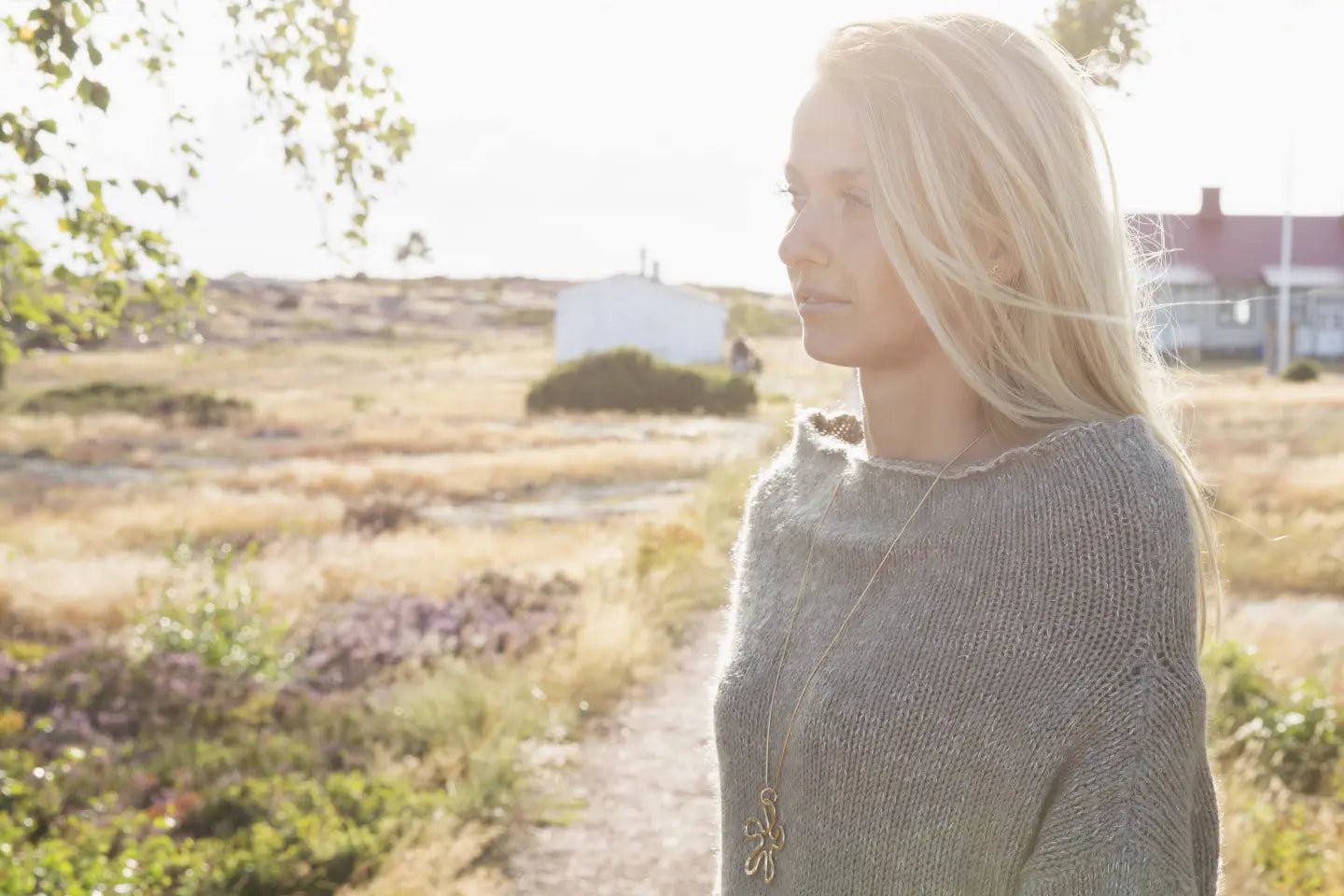 Blonde woman wearing a long gold flower pendant necklace outdoors in warm daylight.