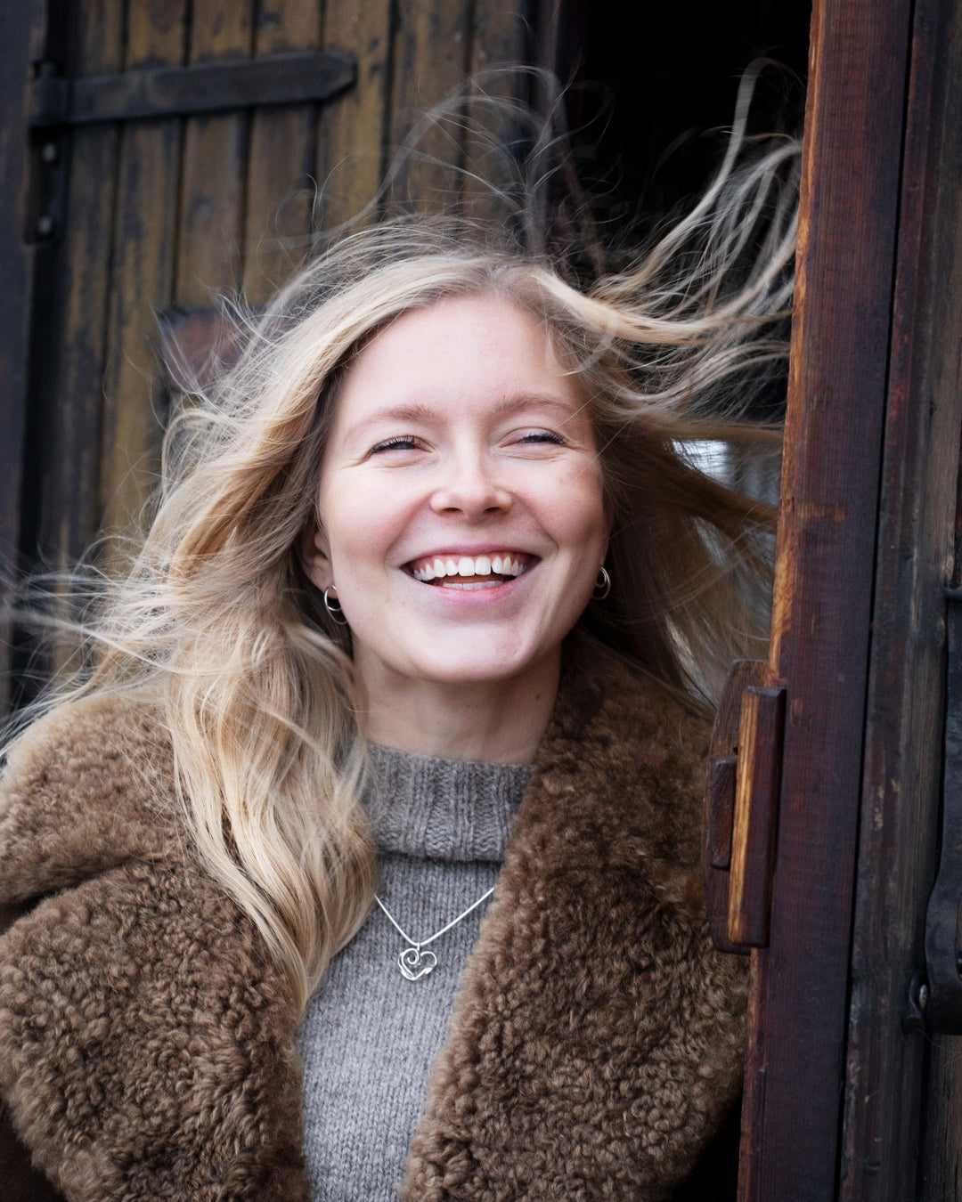 Smiling woman outdoors wearing a silver heart pendant necklace over a gray knit turtleneck and brown shearling coat