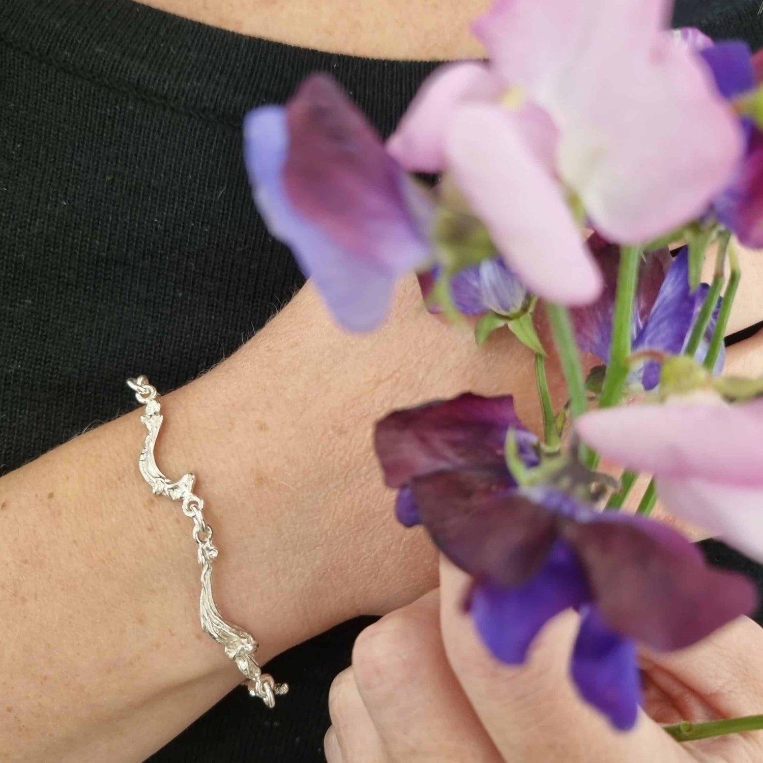 Havet bracelet with wave shaped links worn on the wrist beside purple flowers