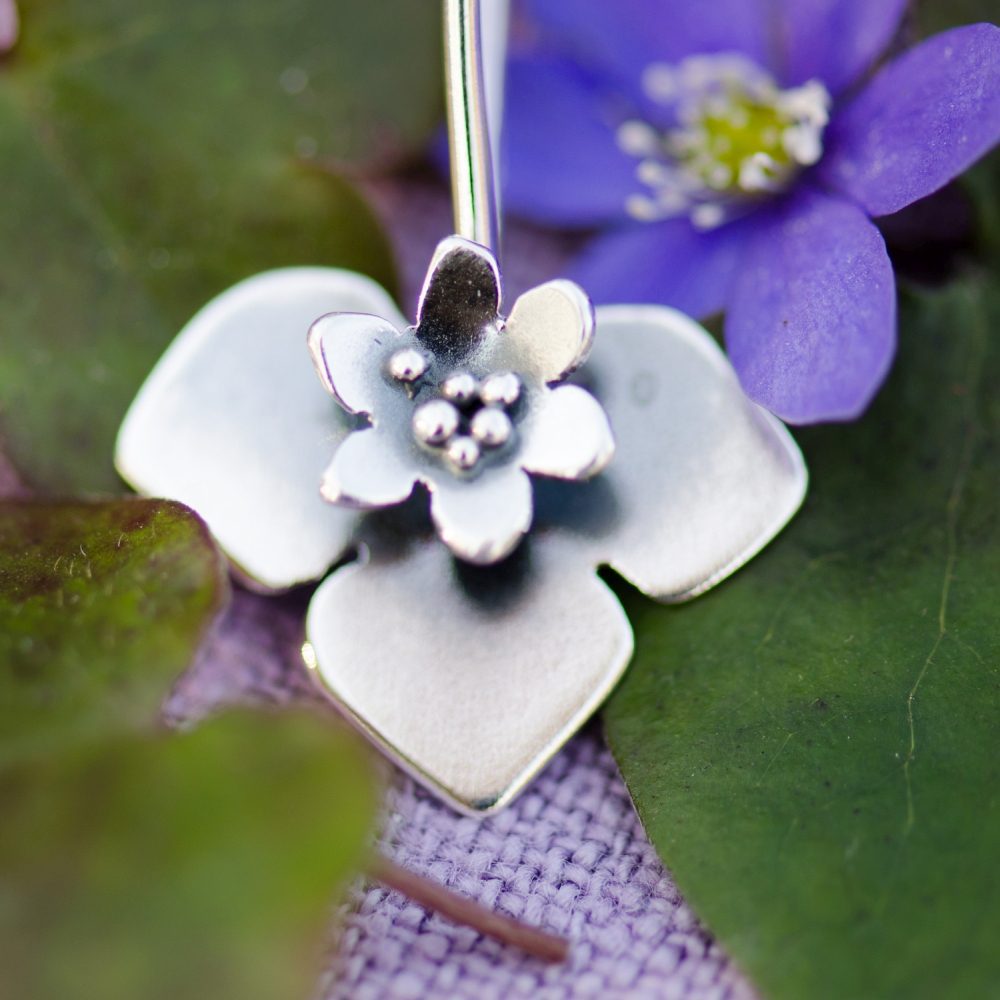 Close-up of the BLÅSIPPA BLAD pendant showing a layered silver flower against a purple bloom and green leaves