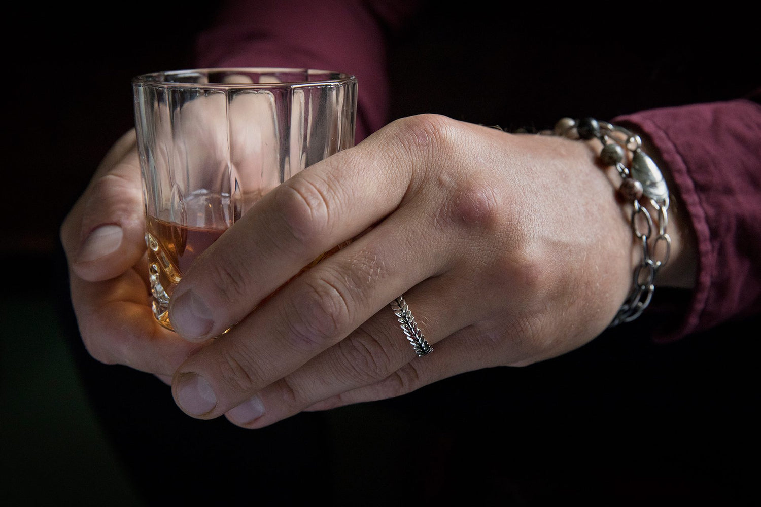 Close up of a hand wearing a narrow silver RÅG ring with a leaf-shaped pattern while holding a glass