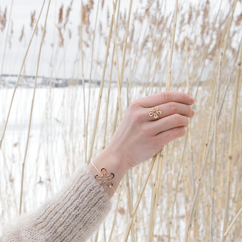 Gold BLOMMAN flower bracelet with a matching ring worn on a hand among tall dried reeds in a snowy scene