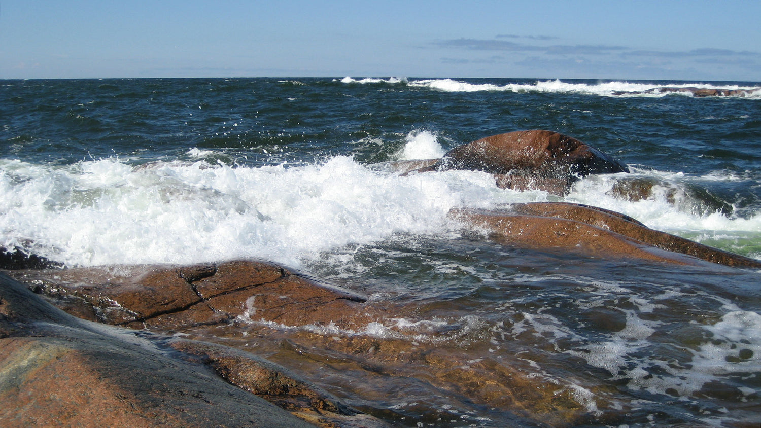 Waves crash against brown rocks along a windswept coast under a bright blue sky.
