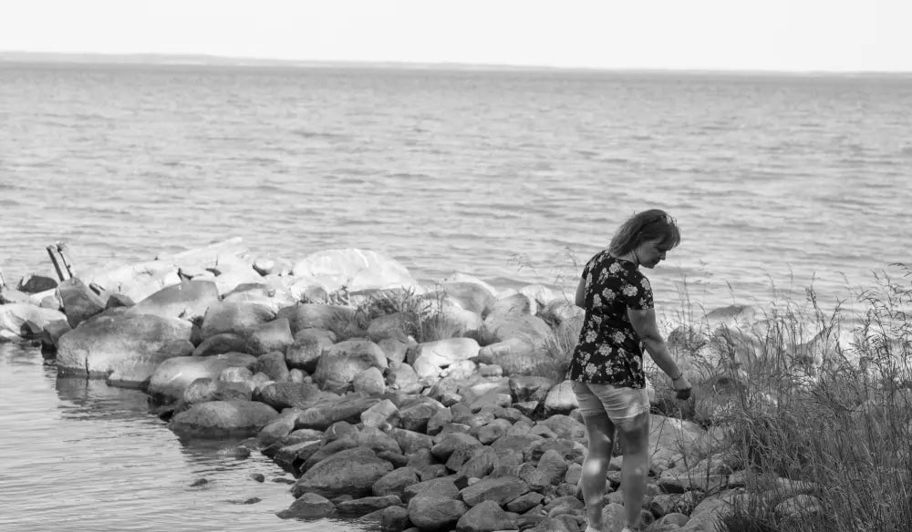 Woman in a floral top and shorts standing on a rocky shore by calm water