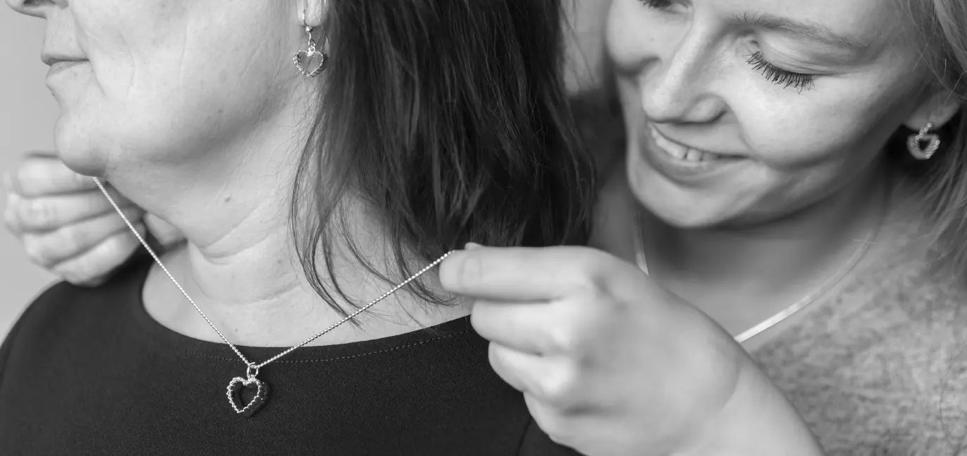 Two women showcase a silver heart shaped pendant necklace in a black and white photograph.