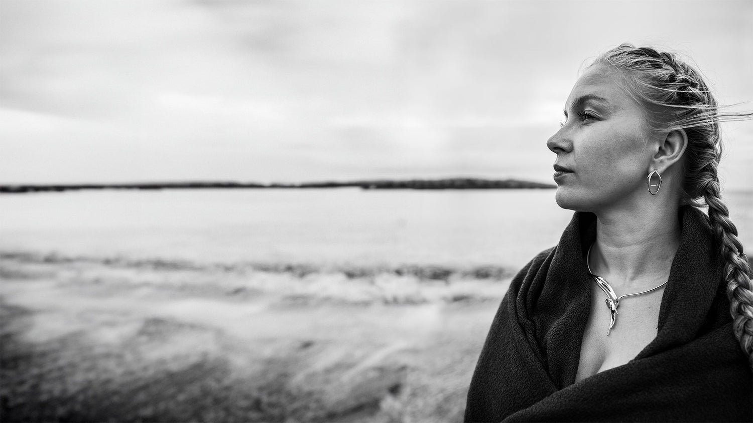Profile portrait of a woman by the sea wearing a dark wrap, braided hair and hoop earrings