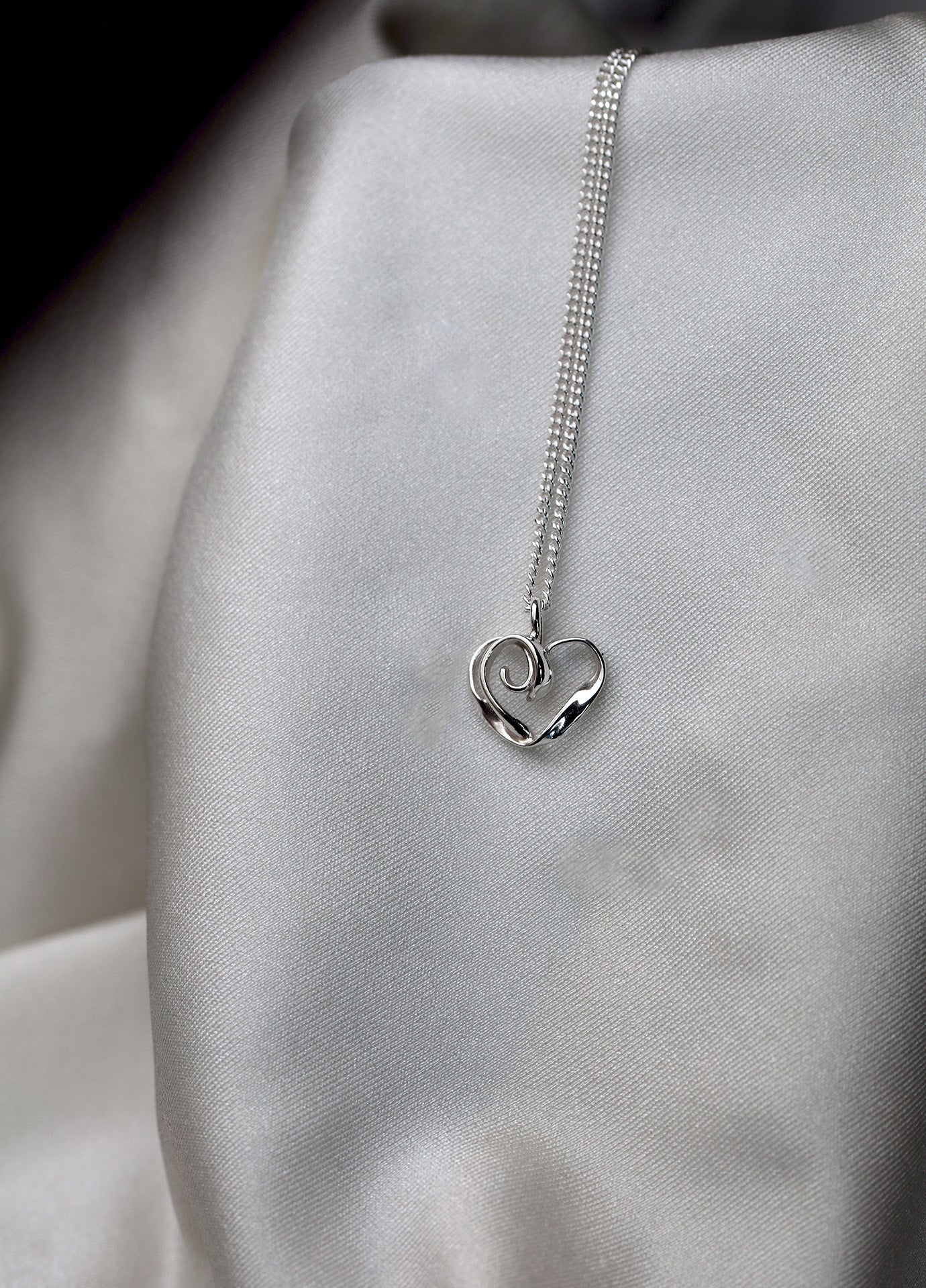 Close-up of a sterling silver chain necklace with a heart pendant on a light fabric backdrop
