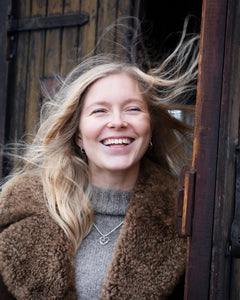 Smiling woman outdoors wearing a silver heart pendant necklace over a gray knit turtleneck and brown shearling coat