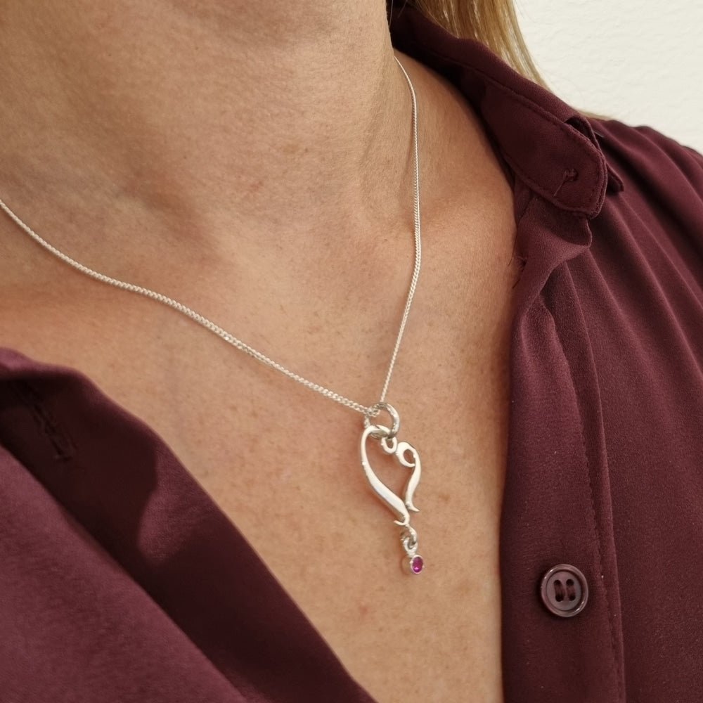 Närbild på silver halsband med stiliserat hjärta och rubinprick nedtill mot huden