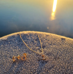 I HAMN LILLA 18K halsband med ring och matchande örhängen i guld liggande på sandig sten i kvällsljus