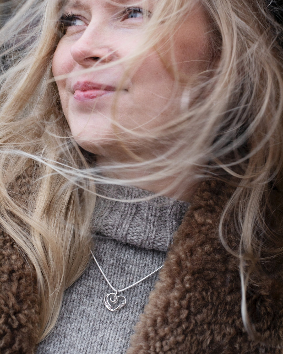 Close-up of a smiling woman wearing the silver heart pendant necklace over a gray knit turtleneck