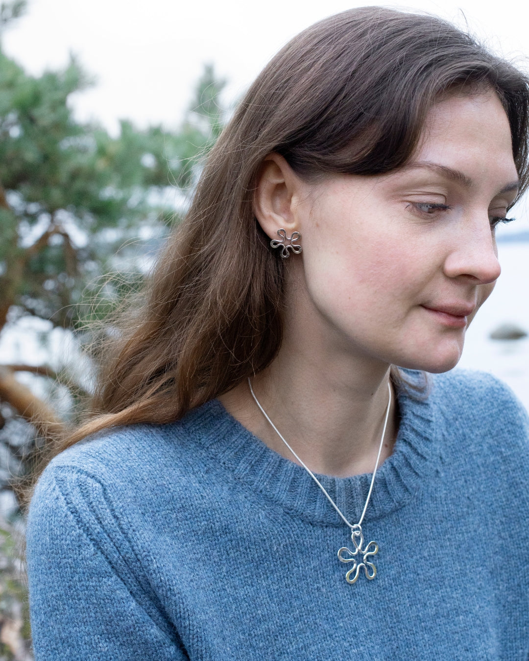 Kvinna i blå tröja bär blomformad silverhalskedja med hänge och matchande örhänge i örat