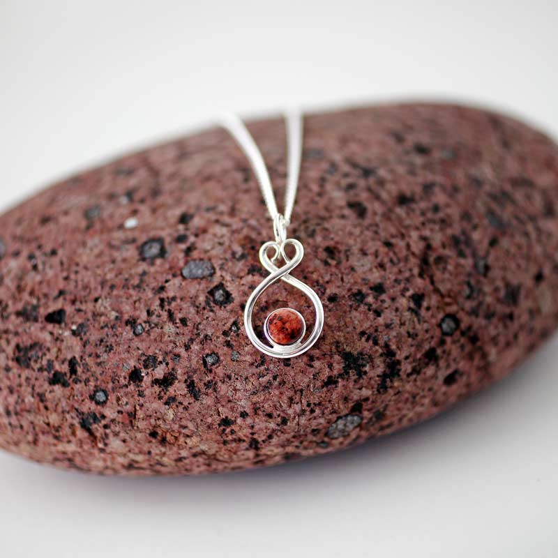 Silver Ålandshjärta halsband med röd granit hängandes framför en röd stenbakgrund