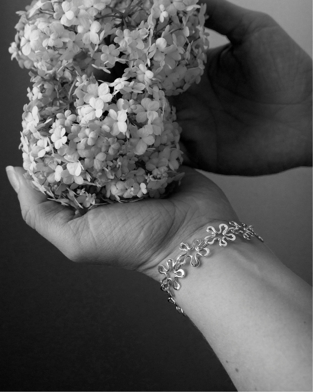 Silver BLOMMAN armband worn on the wrist beside a cluster of small flowers.