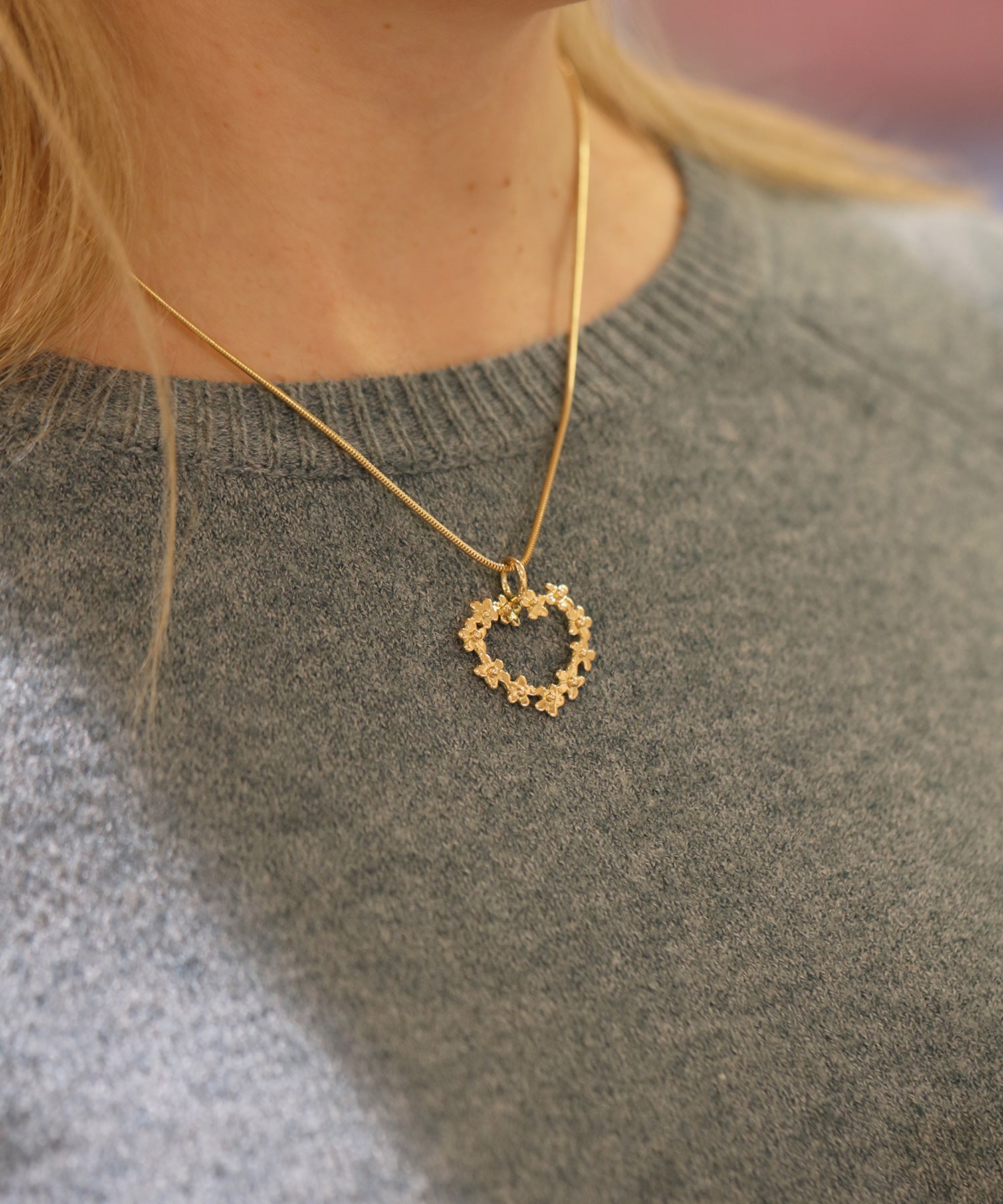 Halsband med blomsterhjärta hänge i guld på tunn kedja bärs av en person i grå tröja
