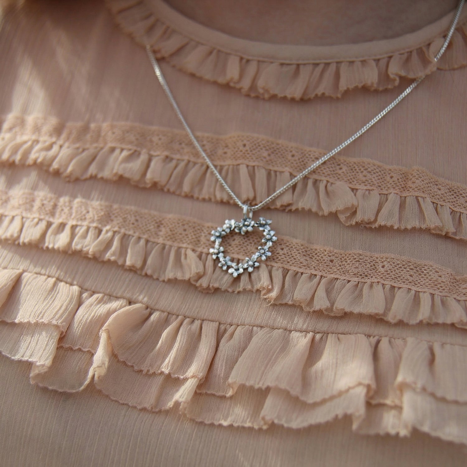 FÖRGÄT MIG EJ HJÄRTA halsband i sterlingsilver med blomformade detaljer mot beige rynkad tygbakgrund.