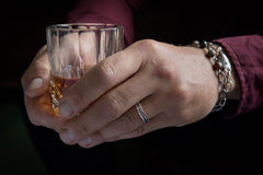 Close up of a hand wearing a narrow silver RÅG ring with a leaf-shaped pattern while holding a glass