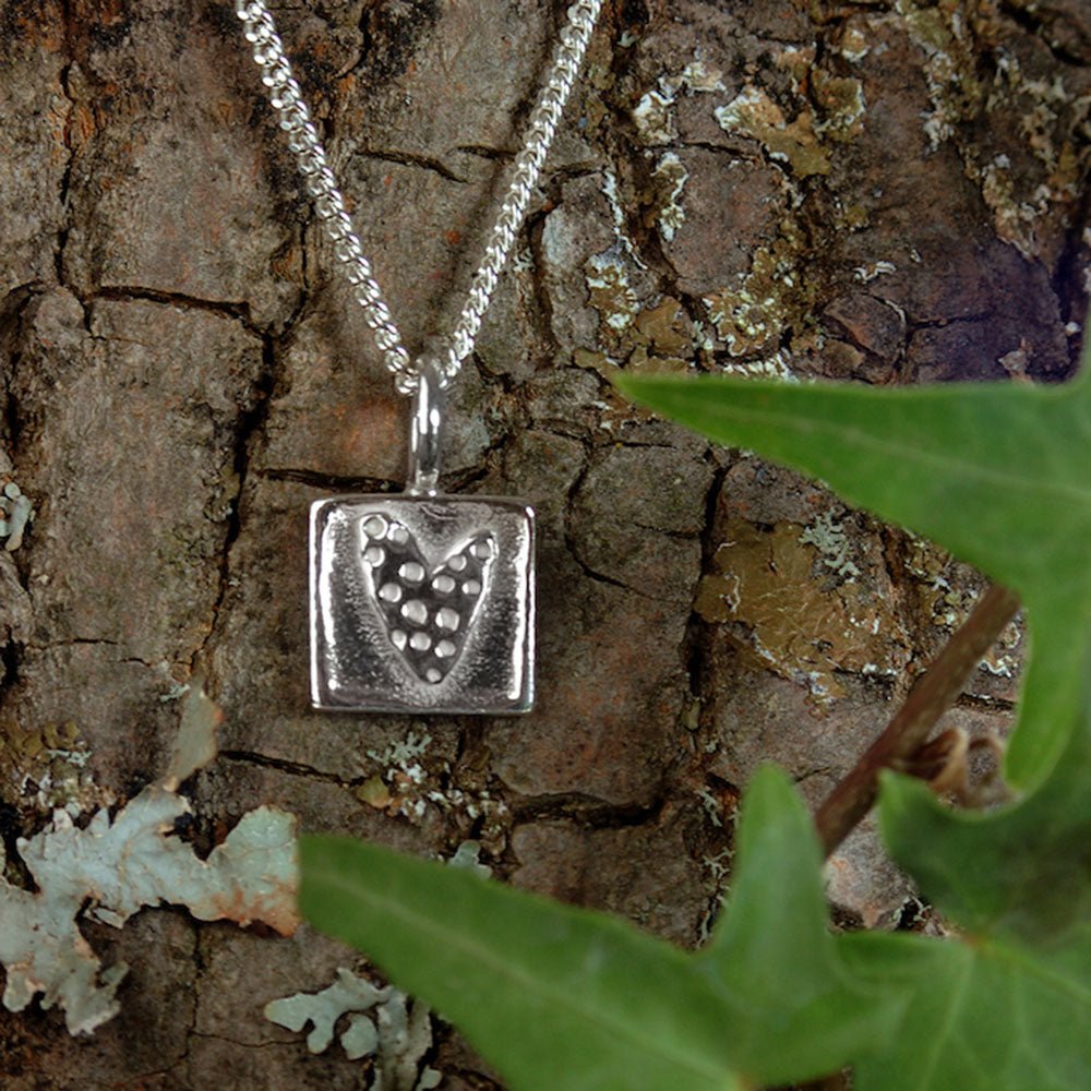 Square silver pendant with a perforated heart design hanging from a silver chain against a tree trunk