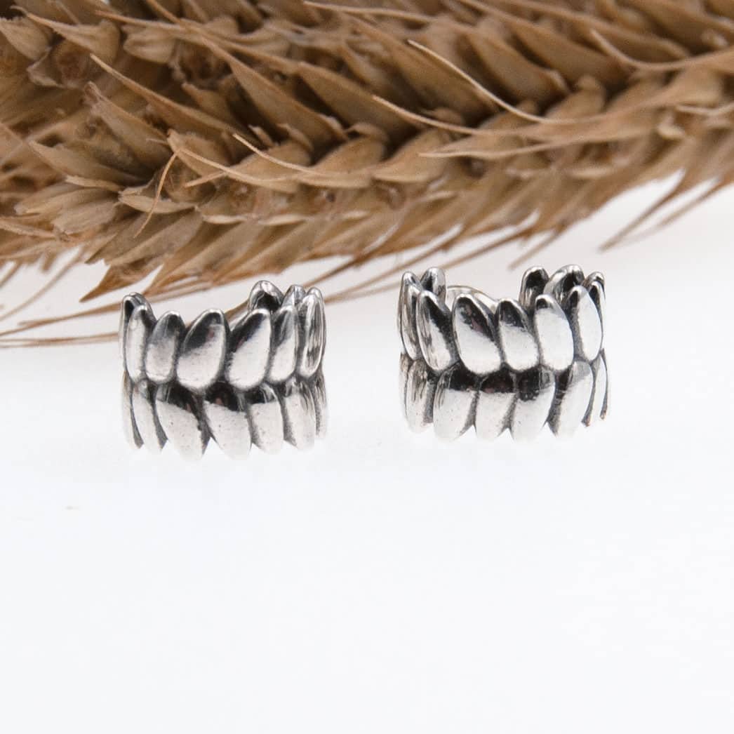 Par RÅG stiftörhängen i 925 sterling silver med bladformade segment ligger bredvid varandra framför torkat gräs