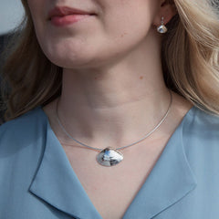 Large polished sandmussla shell pendant on a silver omega chain worn by a woman in a blue blouse.