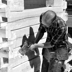 Black and white photo of a craftsman with an axe chopping wood beside stacked timber