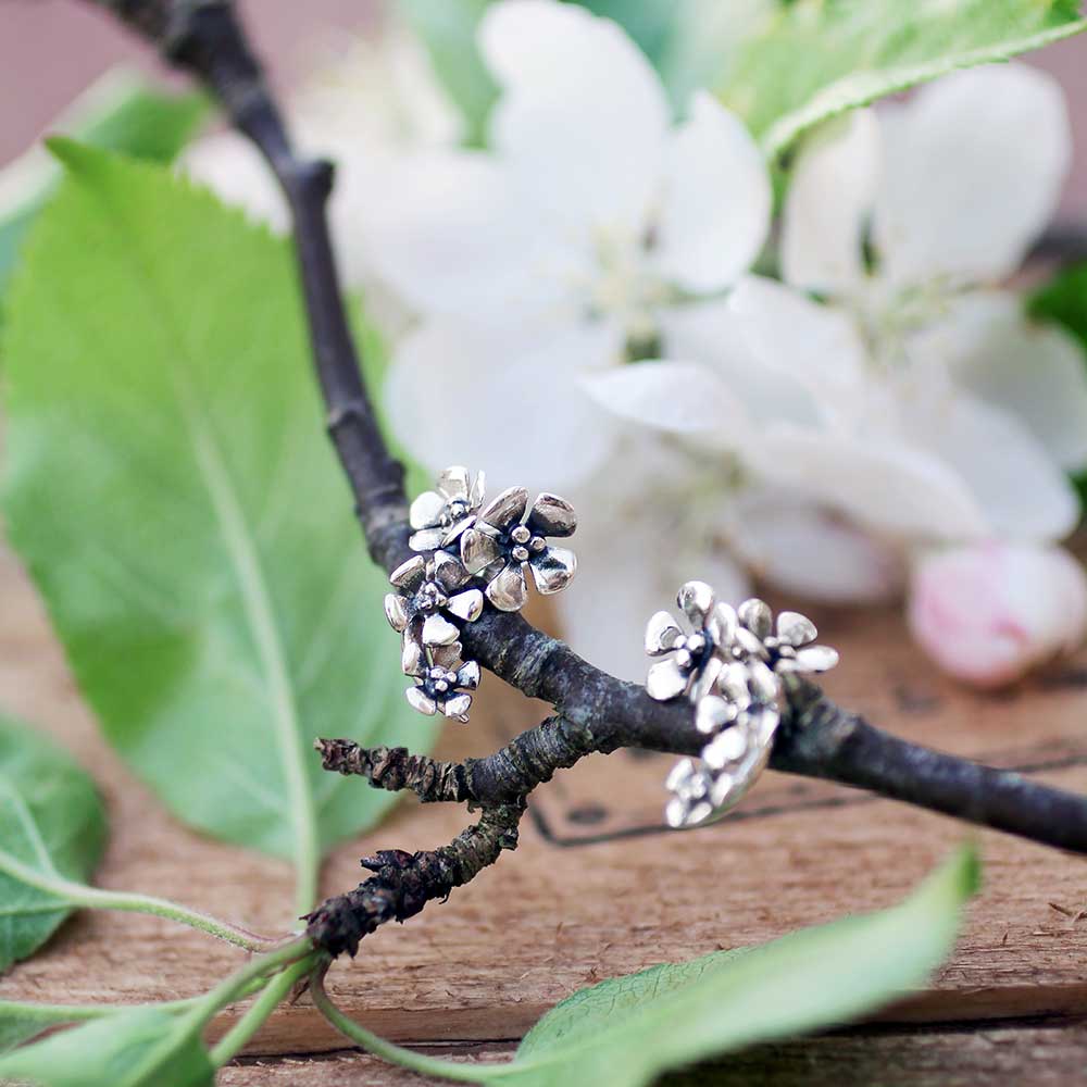 Närbild av ÄPPELBLOM L örhängen på stift i silver på en gren med gröna blad och vita blommor i bakgrunden