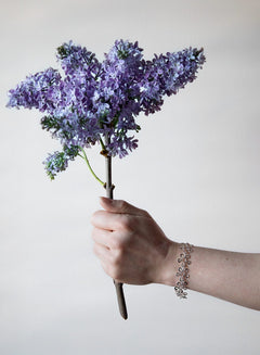 Hand holding a bouquet of purple lilac with a silver BLOMMAN armband on the wrist.
