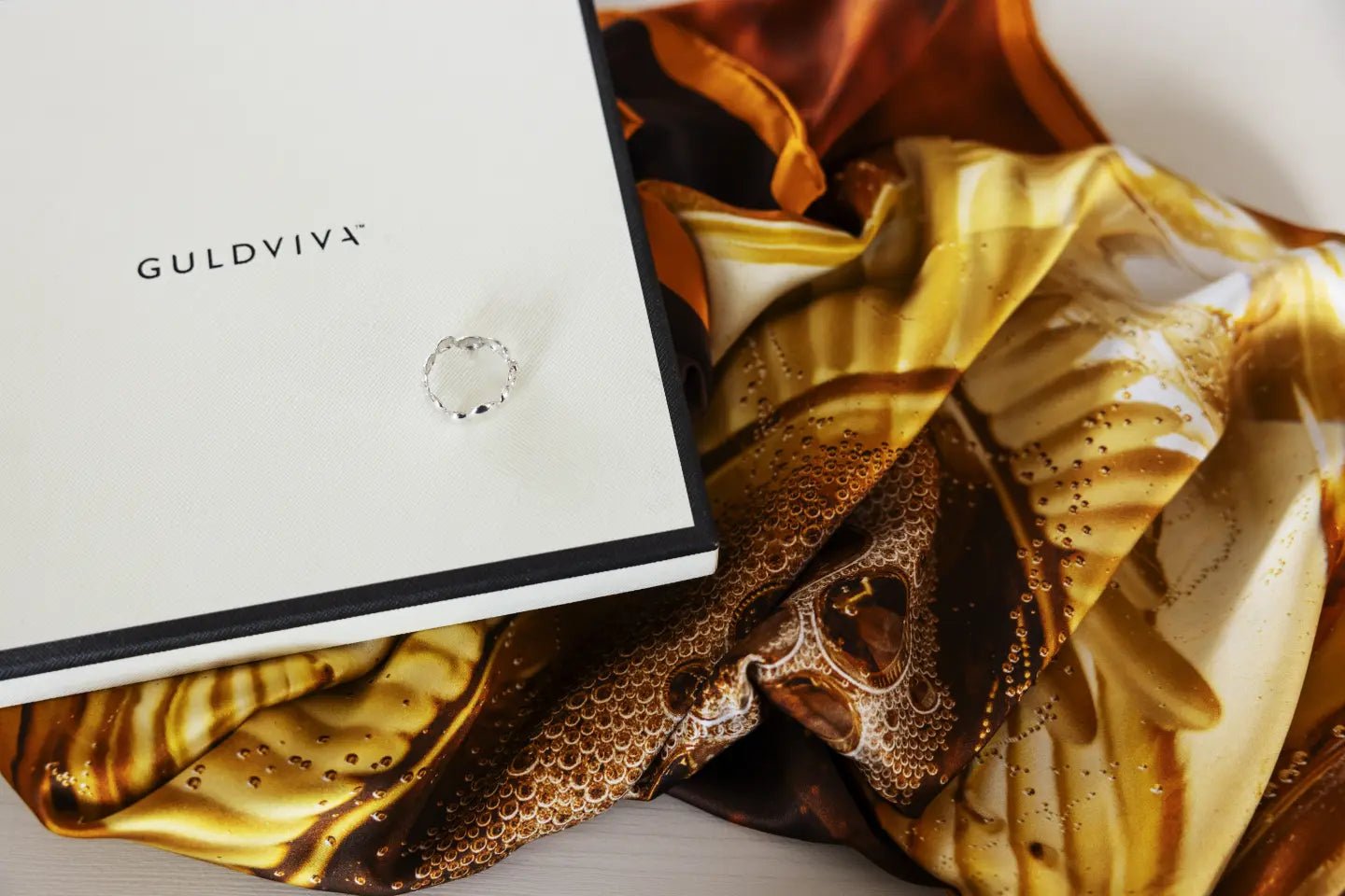 Celebrate Scarfring resting on a white presentation box with a golden patterned scarf.