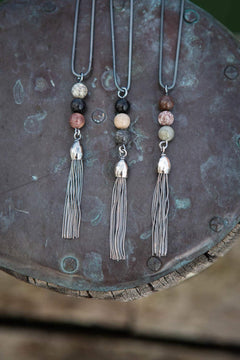 Tre STRAND Fest halsband med färgade strandstenar och grå tofsar hänger mot en metallbakgrund.
