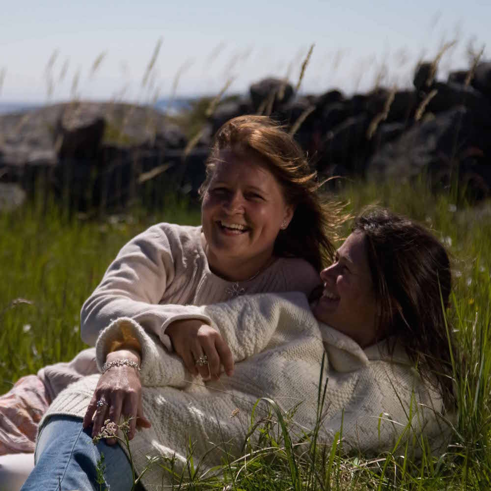 Two friends lying in a grassy field wearing a silver heart pendant necklace.