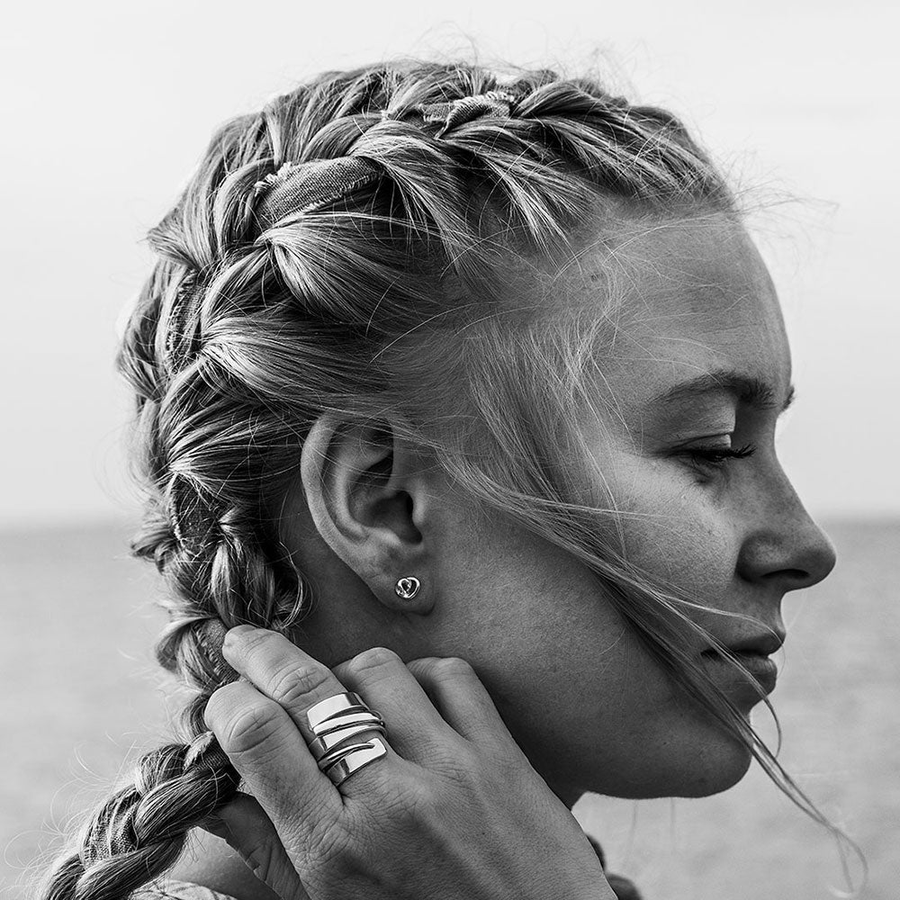 Small heart shaped silver stud earring worn on the ear by a model with braided hair