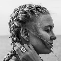 Small heart shaped silver stud earring worn on the ear by a model with braided hair