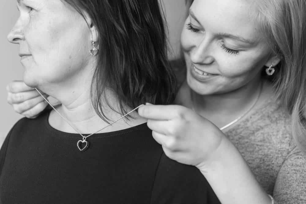Två kvinnor bär ett silverhänge i form av Råghjärta på kedja medan en av dem justerar kedjan runt den andra halsen