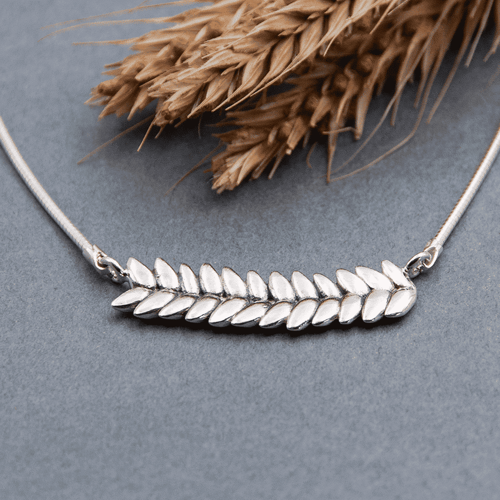 RÅG L necklace in sterling silver with leaf shaped elements on a gray surface with rye stalks in the background