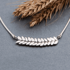 RÅG L necklace in sterling silver with leaf shaped elements on a gray surface with rye stalks in the background